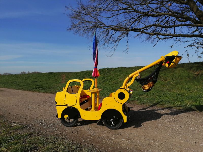 Ein gelber hölzerner Kinderbus als Kinderwagen mit Sonnenschirm steht auf einem Feldweg vor einem grünen Hügel